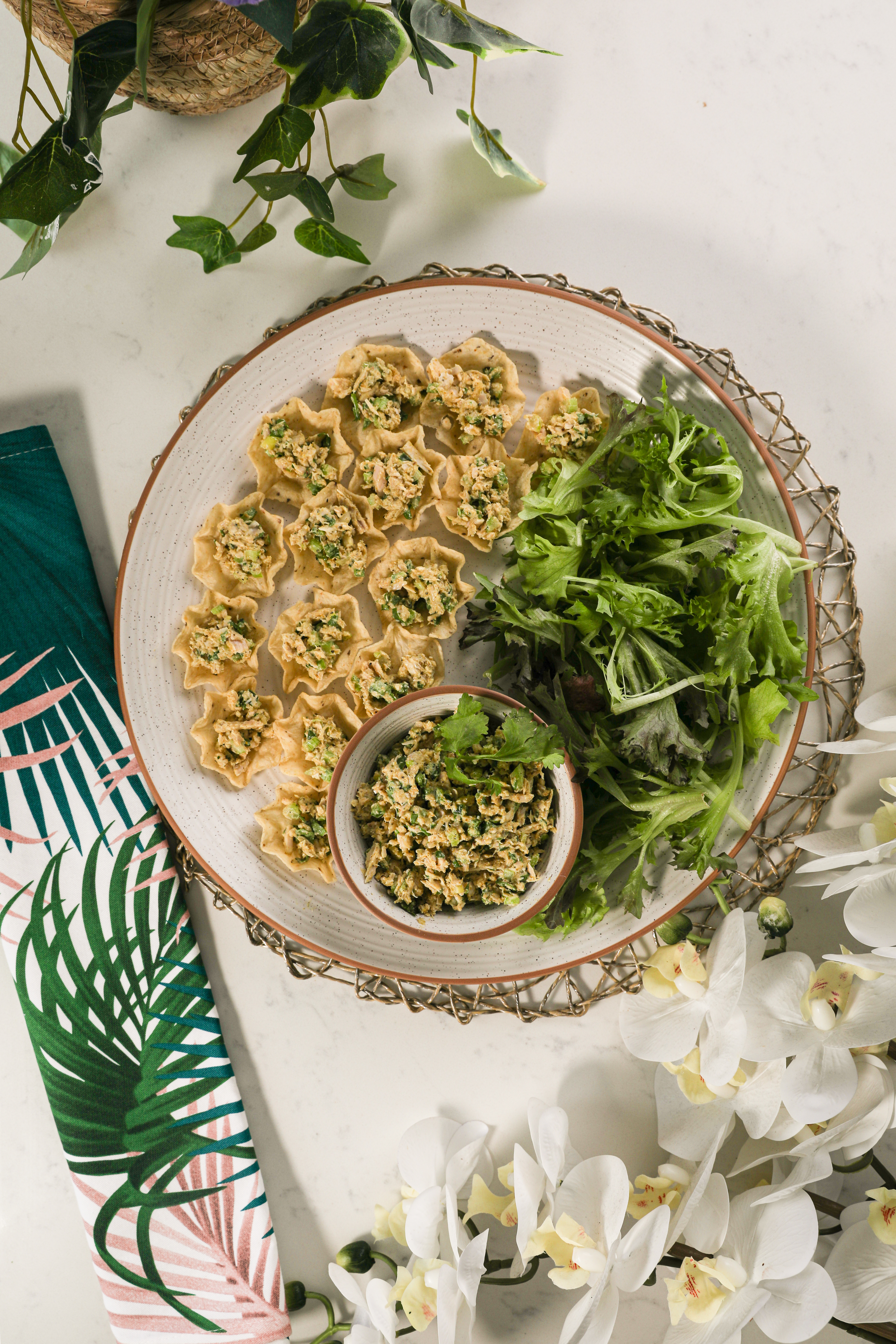 Tortilla scoop chips filled with mashed fish surrounded by greens and a bowl of mashed fish styled with flowers and a printed napkin.