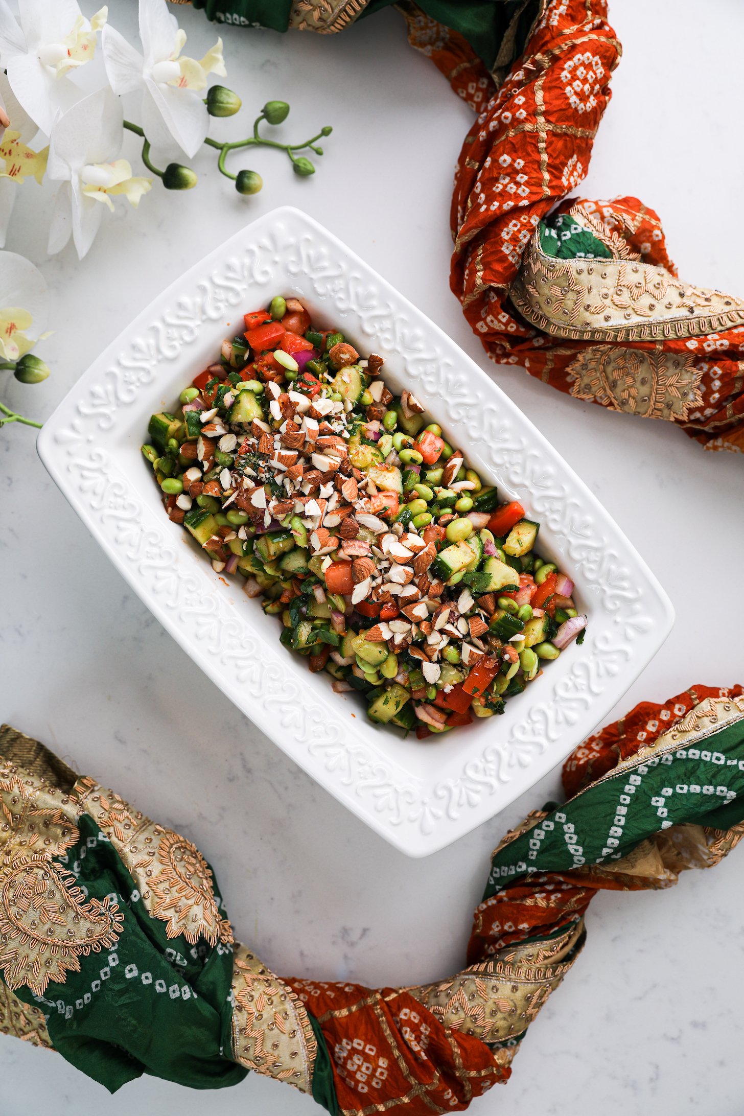 A rectangular bowl filled with chopped vegetables, beans and topped with almonds, styled around a decorative scarf.