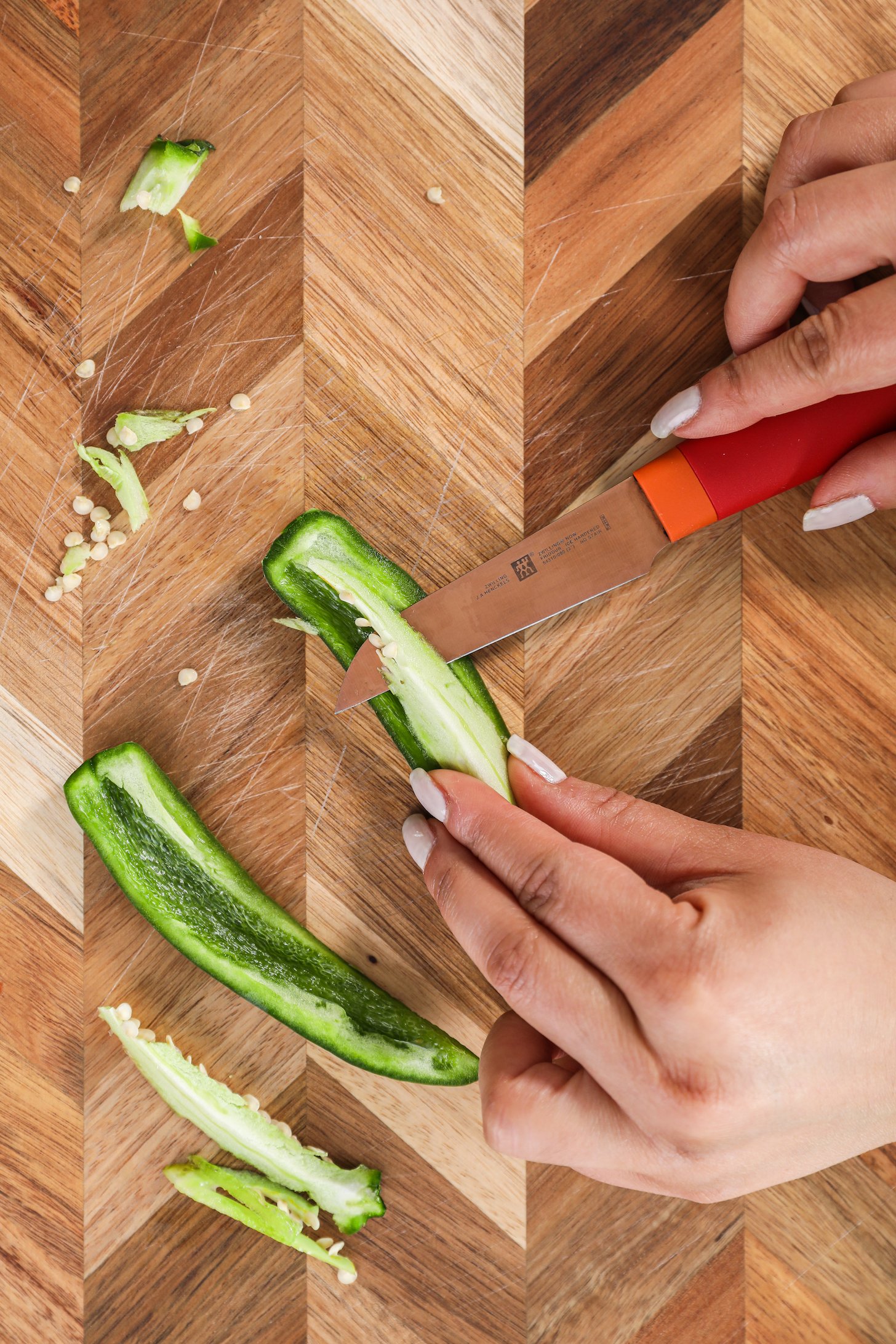 A hand holding a red knife deseeding a green chilli pepper on a wooden board.